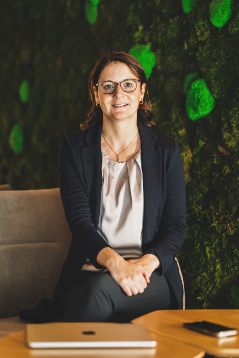 Frau mit Brille und Bluse sitzt an einem Tisch vor einer moosartigen Wand.