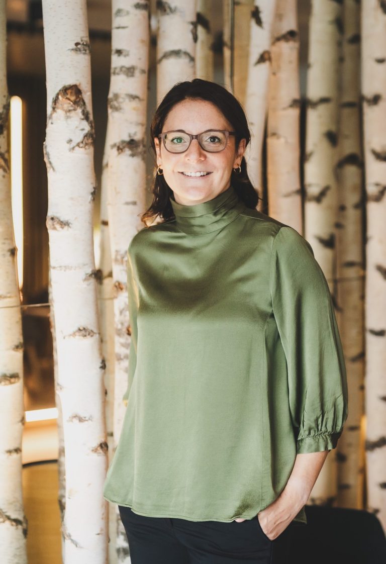Frau mit Brille in bluse aus grünem Stoff vor Birkenstämmen.