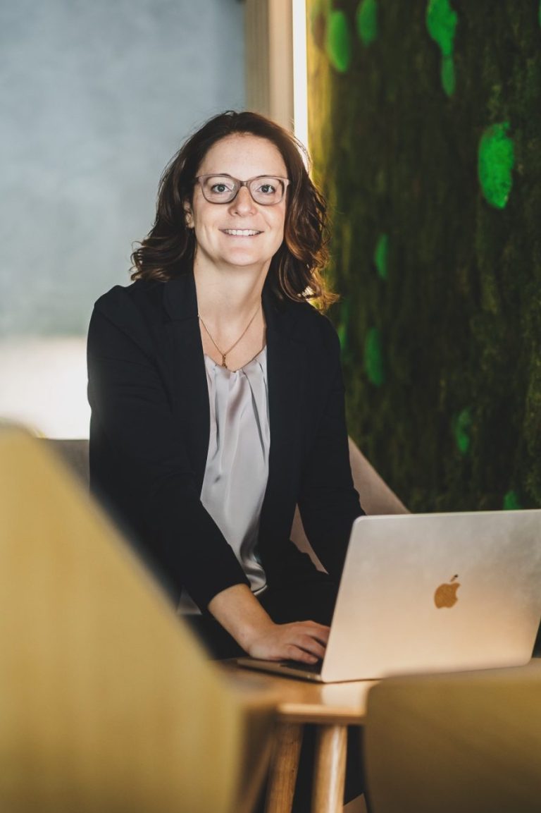 Frau mit Brille und Bluse sitzt an einem Tisch vor einer moosartigen Wand.