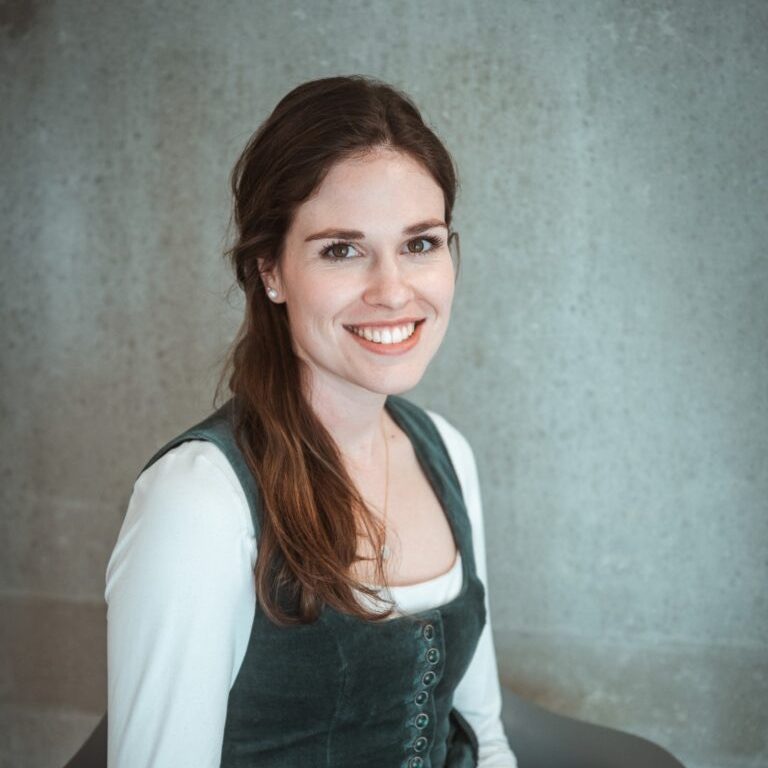 Eine lächelnde Frau mit langen braunen Haaren trägt ein dirndlähnliches Outfit.