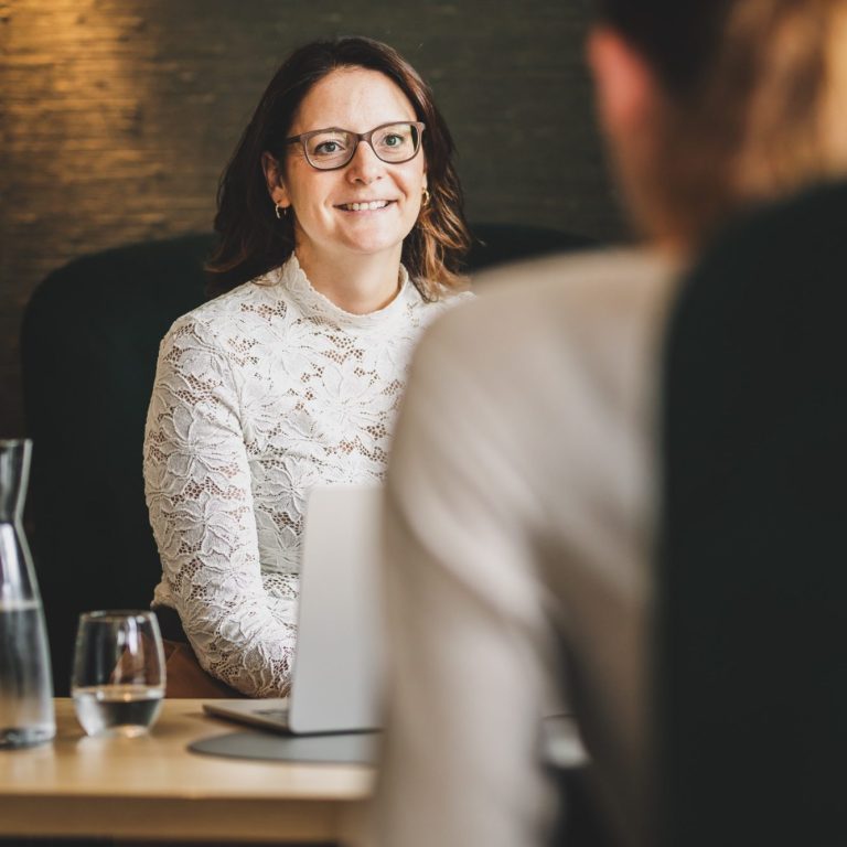 Lächelnde Frau in Brille sitzt an einem Tisch, Blickkontakt mit einer anderen Person.